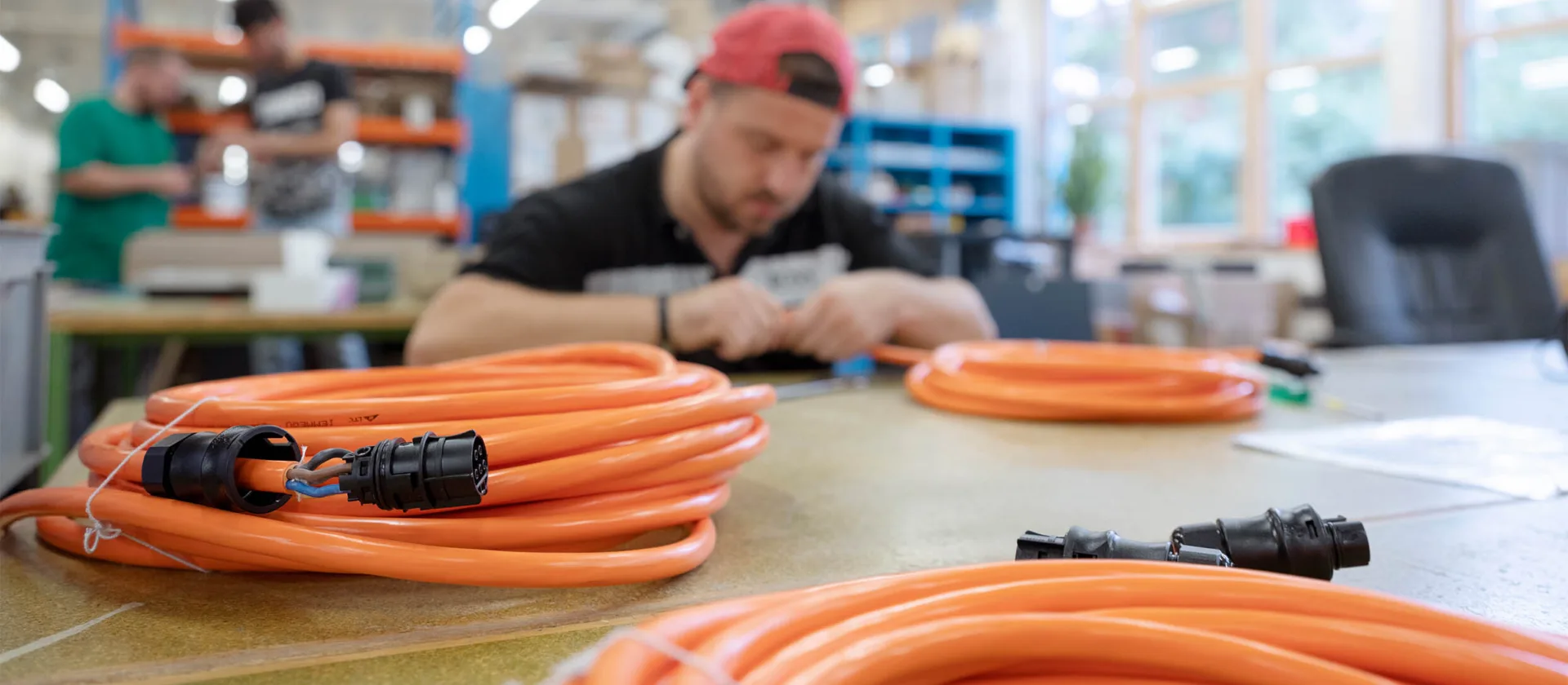Im Vordergrund liegen aufgerollte orange Kabel auf einem Tisch, im Hintergrund sieht man unscharf eine Person, die am Tisch sitzt und arbeitet
